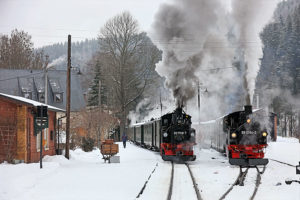 Zwei Dampflokomotiven 99 1568-7 und 99 1594-3 bei der Zugkreuzung im winterlichen Bahnhof mit dampfenden Schornsteinen